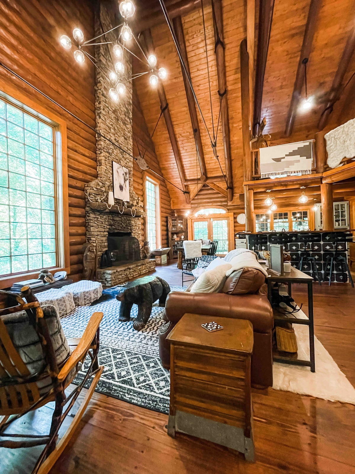 The living area features soaring wooden ceilings and a stack stone fireplace as a focal point. Comfortable seating includes a leather chair and wooden rocking chair, set against a patterned area rug. Natural light filters through large windows, creating a warm and inviting ambiance.