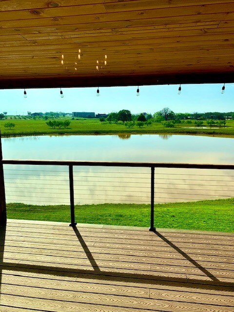A spacious covered balcony provides a view of a serene two-acre tank surrounded by lush green grass. The calm water reflects the sky above, while decorative light bulbs hang from the ceiling, adding a touch of ambiance to the outdoor space.