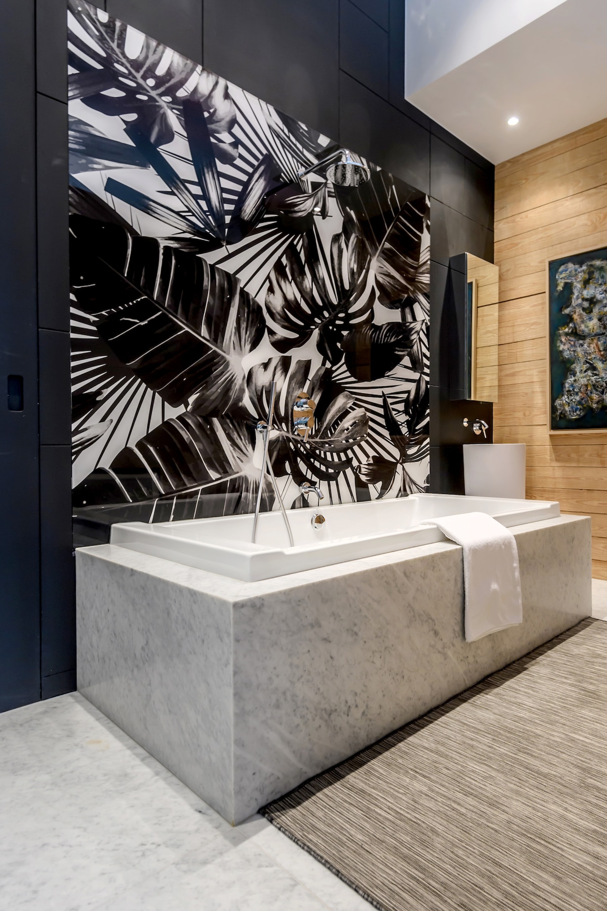 A marble bathtub is positioned against a striking black and white tropical mural. Natural wood accents are present on the walls, complemented by a soft area rug beneath the tub. A modern faucet enhances the sleek design of the bathroom, creating a calming atmosphere.