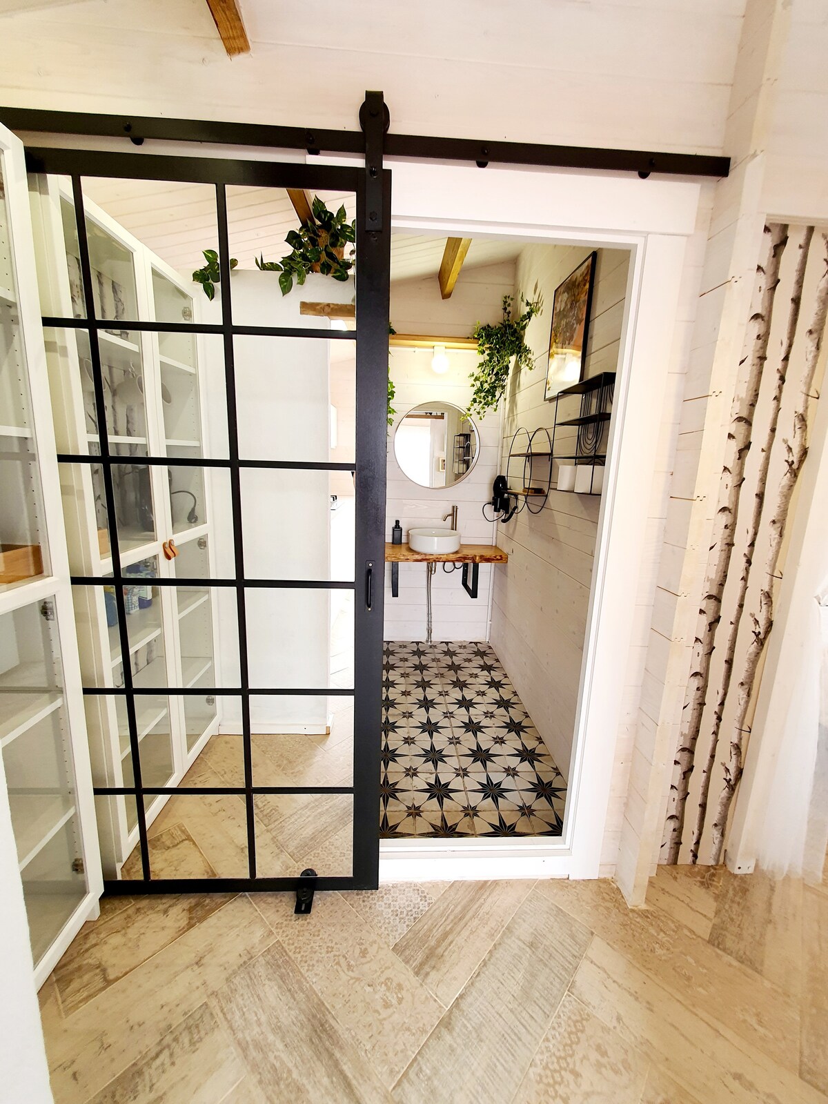 A sliding black barn door leads into the bathroom, framed by white paneled walls. The bathroom features patterned tile flooring, a circular mirror above a wooden sink, and wall-mounted fixtures. Shelving units with glass doors are visible, providing a clean and organized appearance.