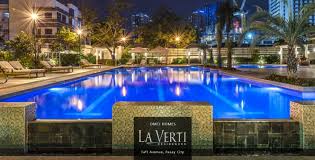 An illuminated swimming pool is surrounded by lounge chairs and greenery, creating a serene outdoor space. The water reflects soft lights from nearby buildings, enhancing the tranquil atmosphere of the area. The La Verti sign is prominently displayed near the pool.