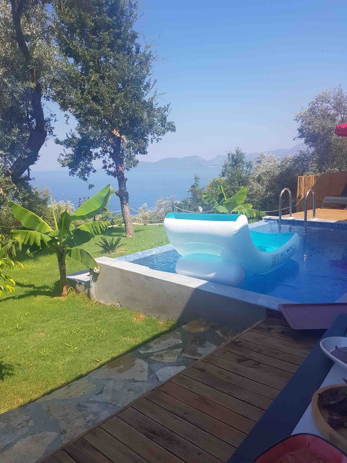 A serene outdoor area features a swimming pool with a floating lounge chair, surrounded by lush greenery and tropical plants. In the background, a calming view of the water and distant hills is visible under a clear blue sky.