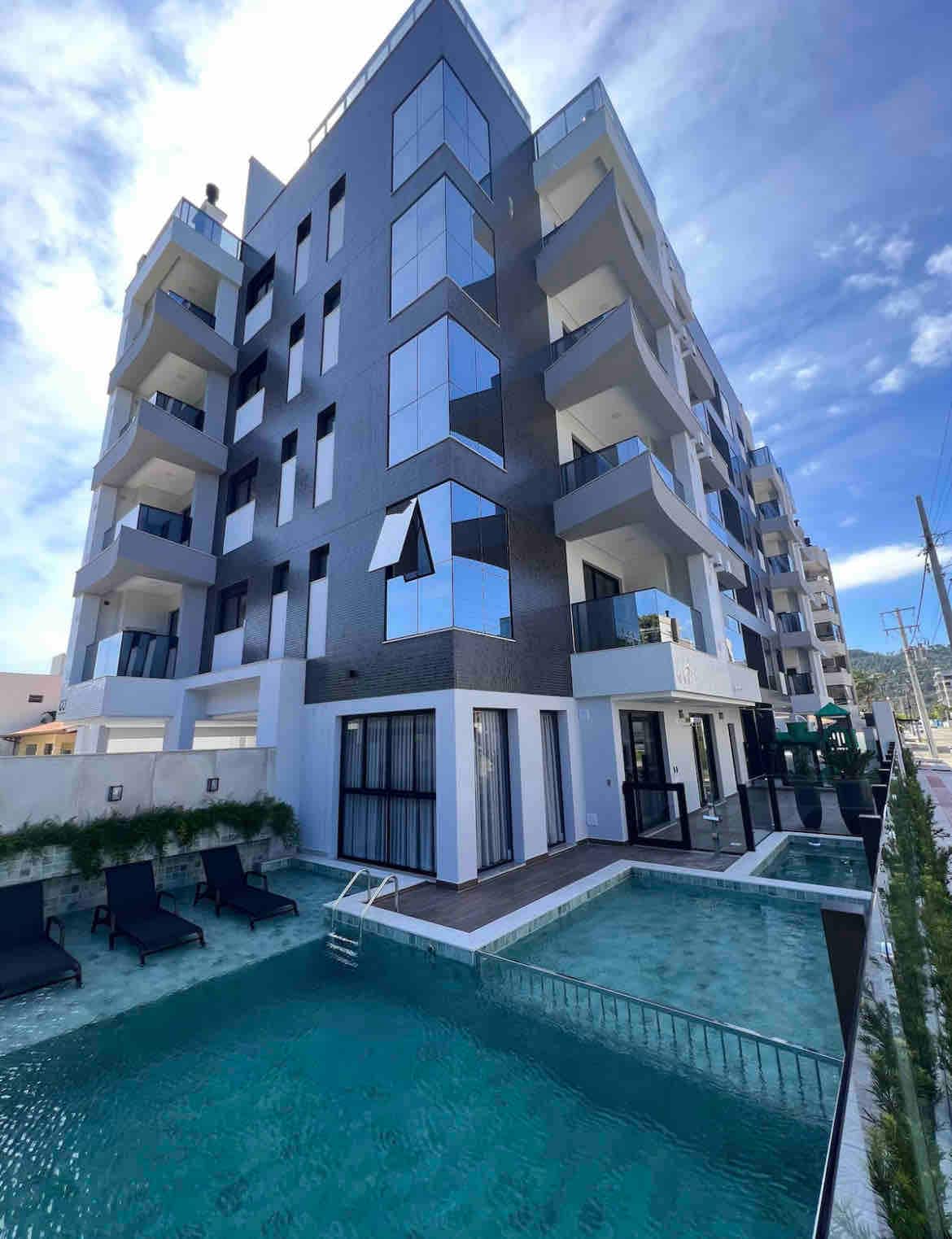 A modern multi-story building showcases large glass windows and balconies. A sleek swimming pool is visible at the base, surrounded by lounge chairs and landscaped areas, reflecting the clear blue sky above and creating a serene atmosphere for guests.