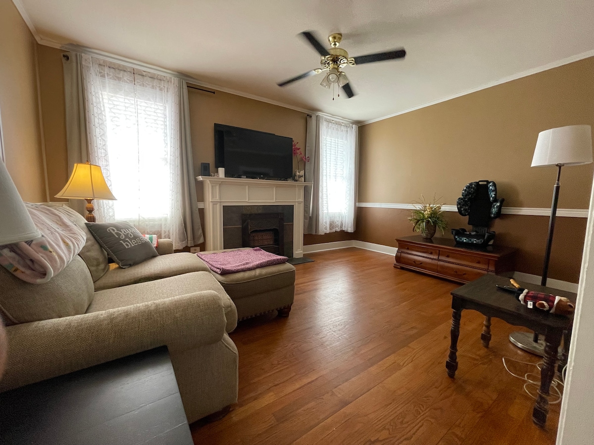 A cozy living room features a light-colored sofa with a decorative pillow. A wooden coffee table is positioned nearby, and a fan hangs from the ceiling. A television is mounted above a fireplace, with windows dressed in light curtains allowing natural light to fill the space.