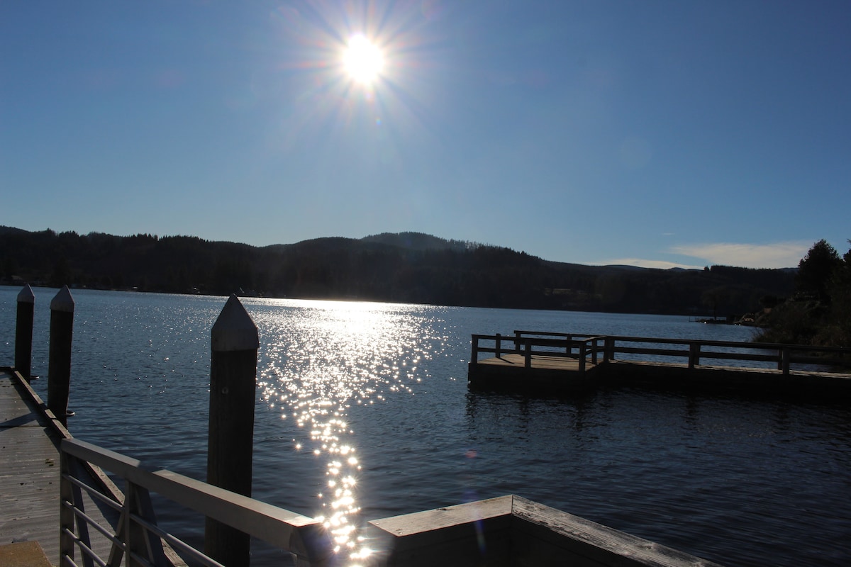A serene view of a sparkling lake reflects sunlight under a clear blue sky. Pier posts line the edge, leading towards a wooden dock in the distance amidst gently rolling hills, creating a peaceful atmosphere.