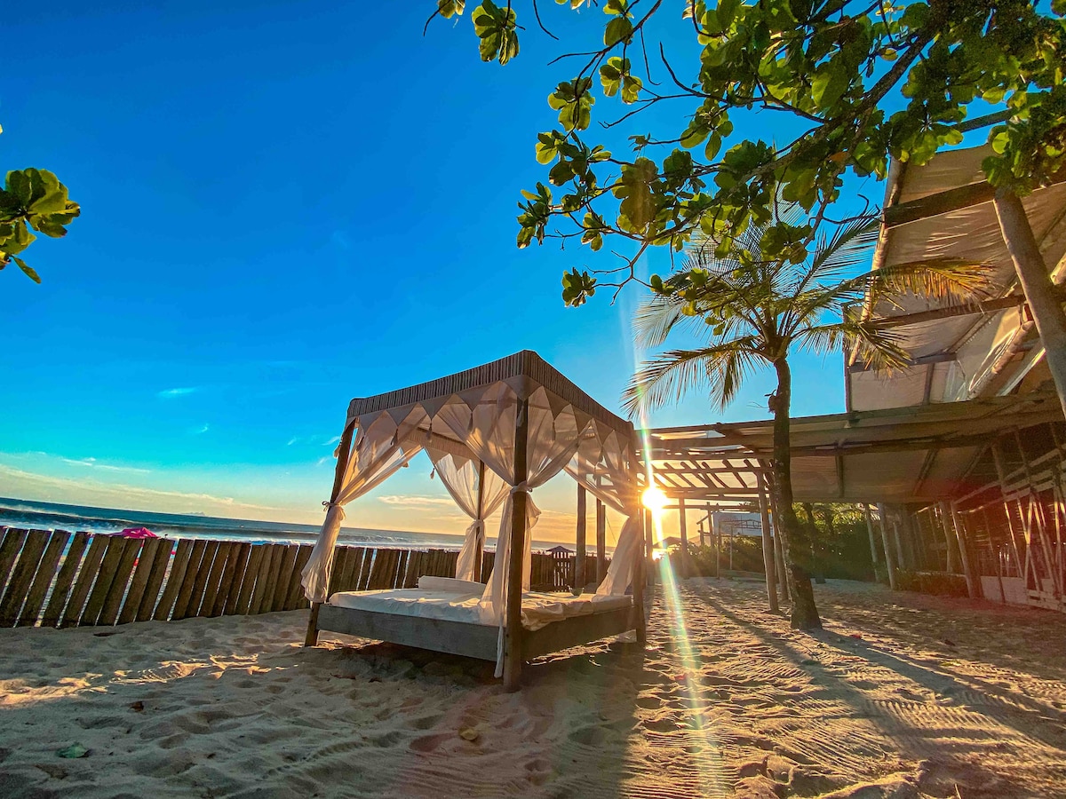 A beachfront cabana is positioned on golden sand, framed by lush palm trees. The rising sun casts a warm glow, creating a serene atmosphere. The shaded cabana is draped with light fabric, inviting relaxation and tranquility with beach views extending beyond.