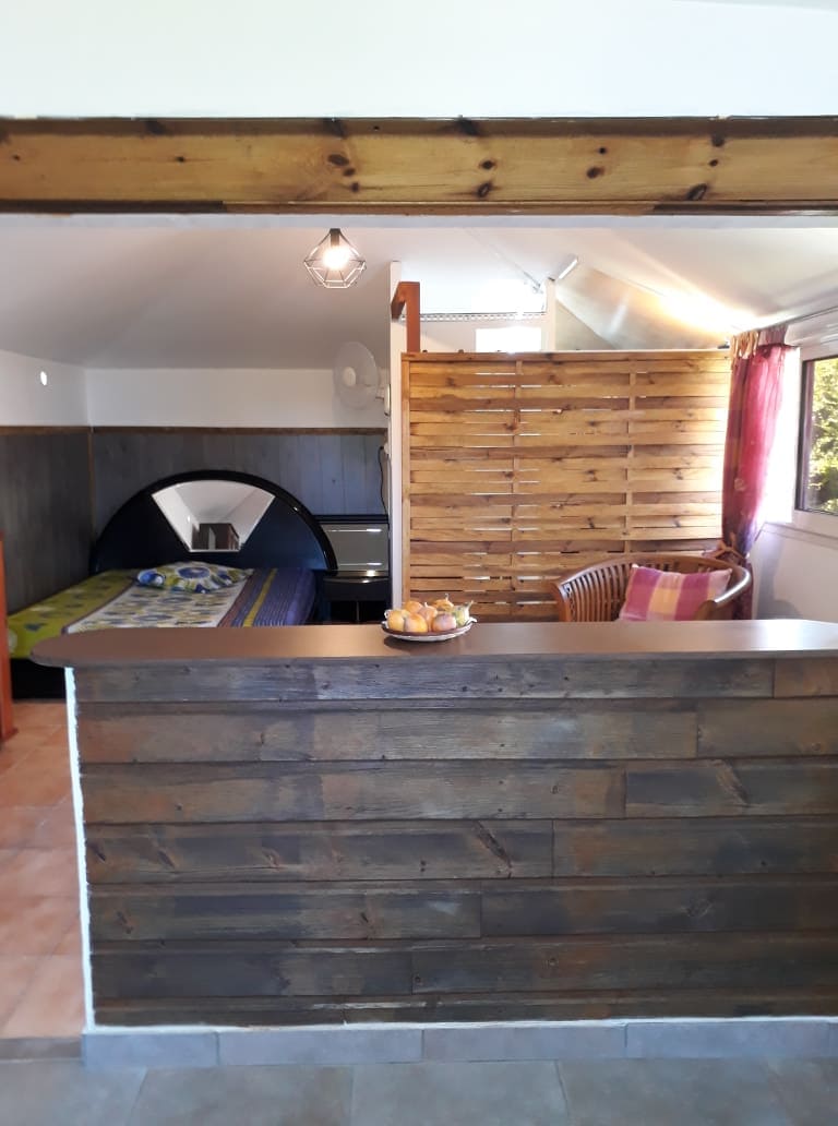 An open studio layout is presented, featuring a wooden reception area with a counter. A cozy seating area includes a wicker chair adorned with decorative pillows. The sleeping space is visible, highlighted by soft lighting and a patterned bedspread, creating a warm and functional environment.