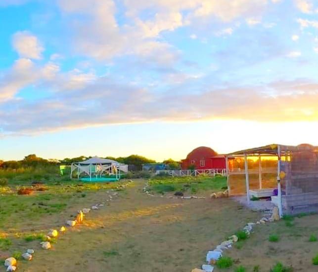 An outdoor area is depicted with a sandy path lined by stones, leading towards several structures. Tents and cabins can be seen against a backdrop of a setting sun, providing a warm golden hue. Expansive fields and scattered greenery are visible throughout the landscape.