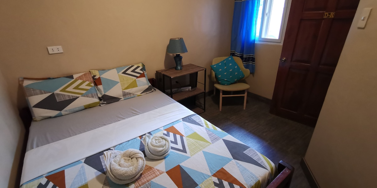 A cozy room is presented with a snug double bed adorned in colorful geometric patterns. A small table with a lamp and a comfortable chair are positioned nearby. Soft lighting streams through the window dressed in blue curtains, enhancing the welcoming ambiance.