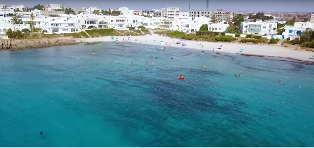The image captures a serene beach scene with clear turquoise water gently lapping at the shore. Families and individuals are seen enjoying the water, with small colorful umbrellas providing shade on the sandy beach. A backdrop of modern white buildings complements the coastal landscape.
