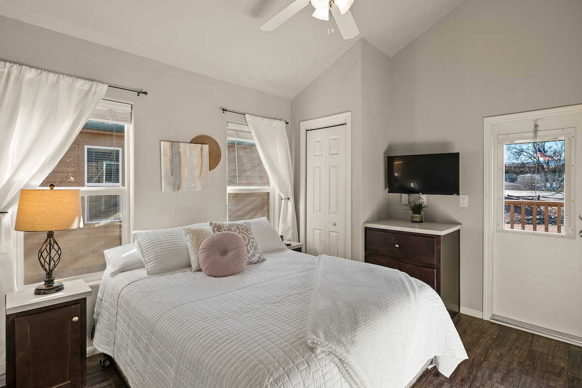 A well-lit bedroom is presented with a spacious bed covered in a white quilt, accentuated by decorative pillows. Windows frame the room, allowing natural light to flood in. A dresser and a mounted TV are positioned against the wall, creating a cozy and functional retreat.