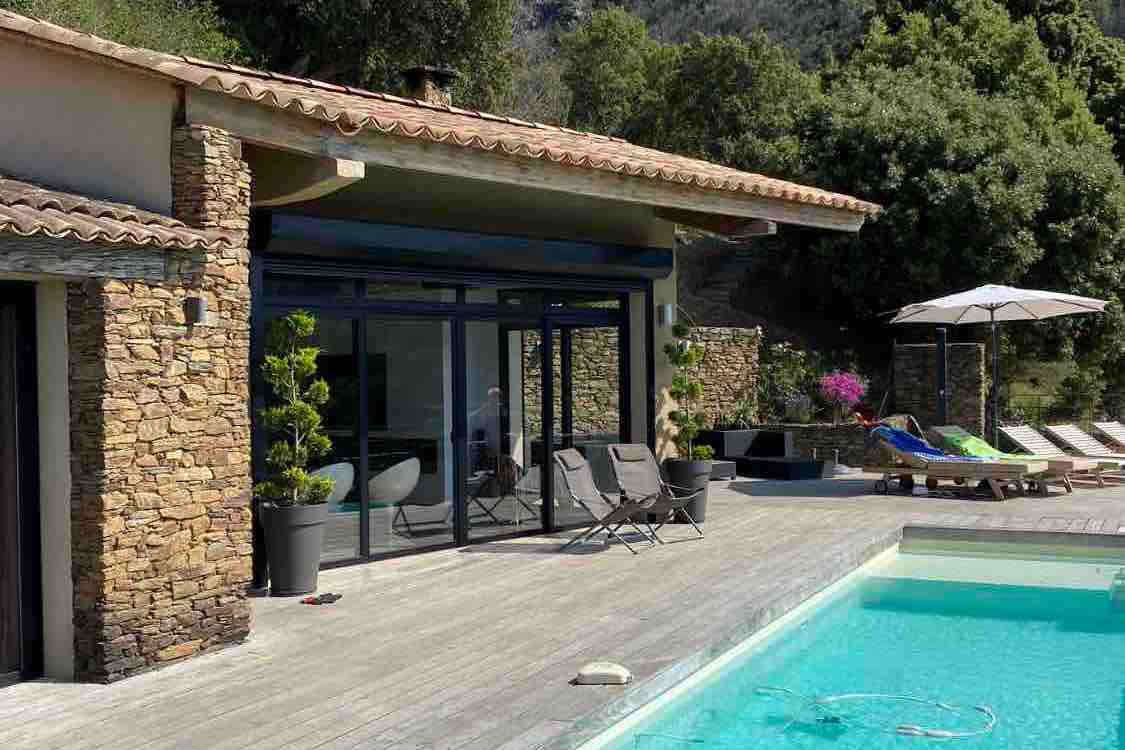 A modern villa exterior is shown, featuring large glass doors that open to a spacious wooden deck with sun loungers and an umbrella. A tranquil infinity pool reflects the surrounding greenery. The stone walls add a natural, rustic touch to the contemporary design.