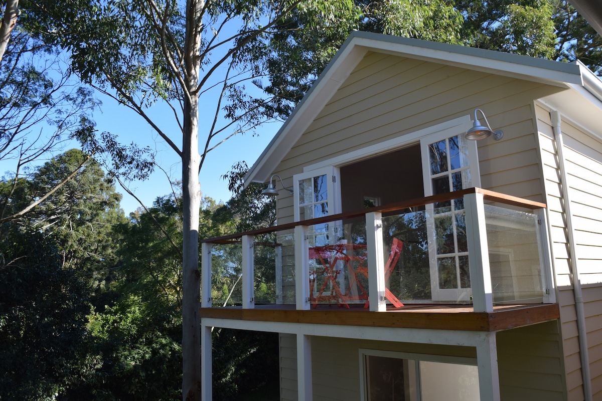 The exterior of the Airbnb showcases a charming two-story structure with a balcony. Glass railing provides a clear view of the surrounding greenery. Large windows allow natural light to fill the interior, while tall trees accentuate the peaceful country setting.