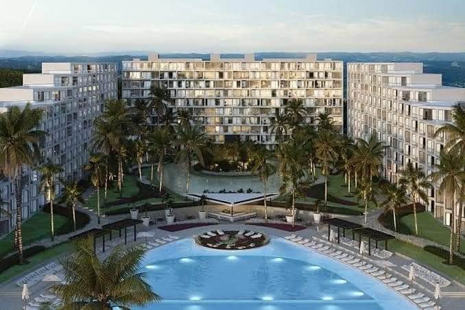 A modern resort complex is surrounded by lush palm trees and manicured lawns. A large swimming pool can be seen at the forefront, with lounge chairs arranged neatly around it. The multi-story building features numerous balconies and large windows, reflecting the bright sky.