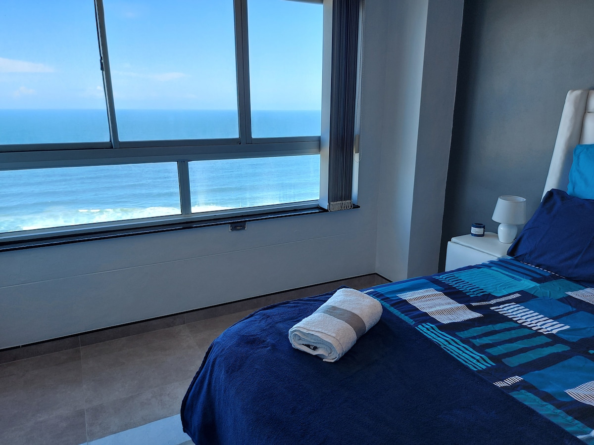 A bright bedroom space features a spacious bed dressed in blue and patterned bedding. A window offers expansive views of the sea, while a bedside table displays a lamp. A neatly folded towel is placed on the bed, enhancing the inviting atmosphere.