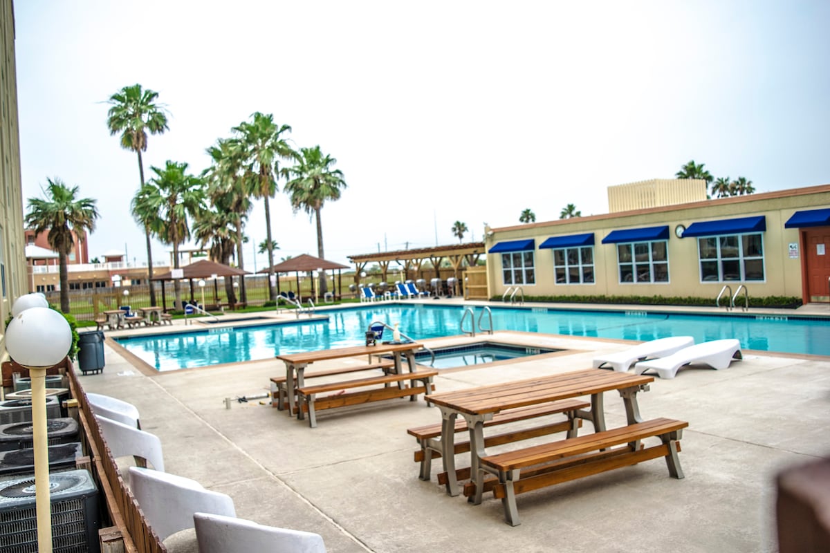 An expansive swimming pool is surrounded by palm trees, with lounge chairs and shaded areas for relaxation. Several wooden picnic tables are positioned nearby, offering ample space for outdoor dining. The serene atmosphere is complemented by well-maintained landscaping.