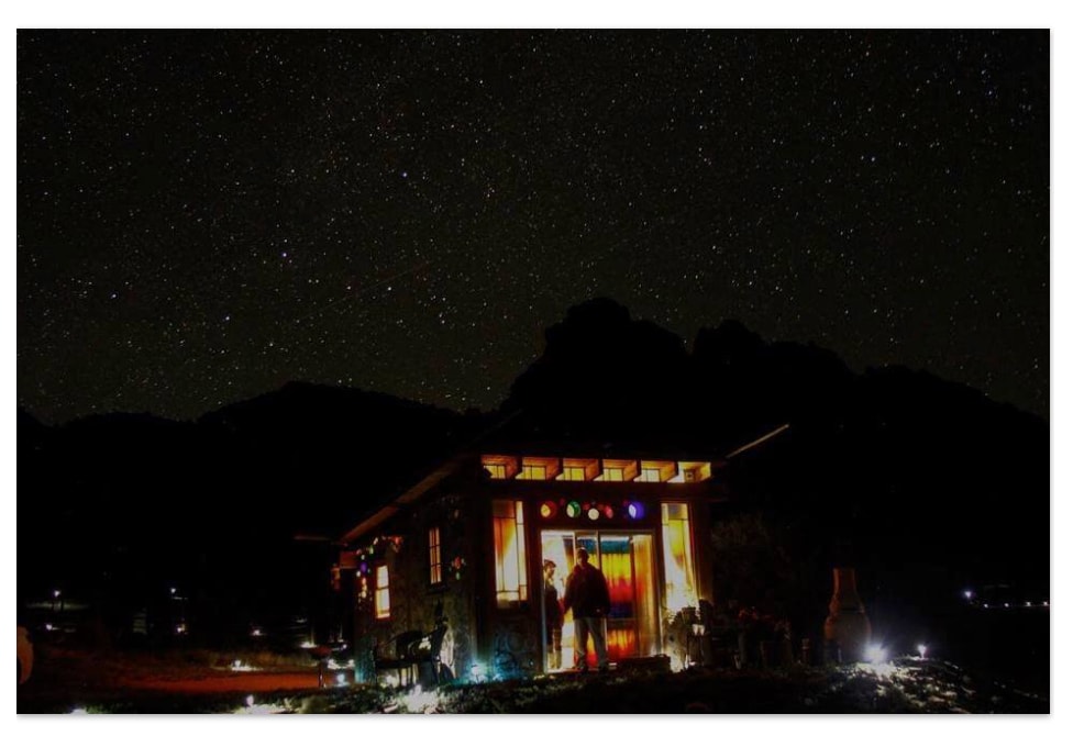 A cozy cottage is illuminated warmly against a backdrop of a star-filled night sky. The silhouette of surrounding mountains adds depth to the scene, while decorative lights enhance the cottage's inviting exterior.