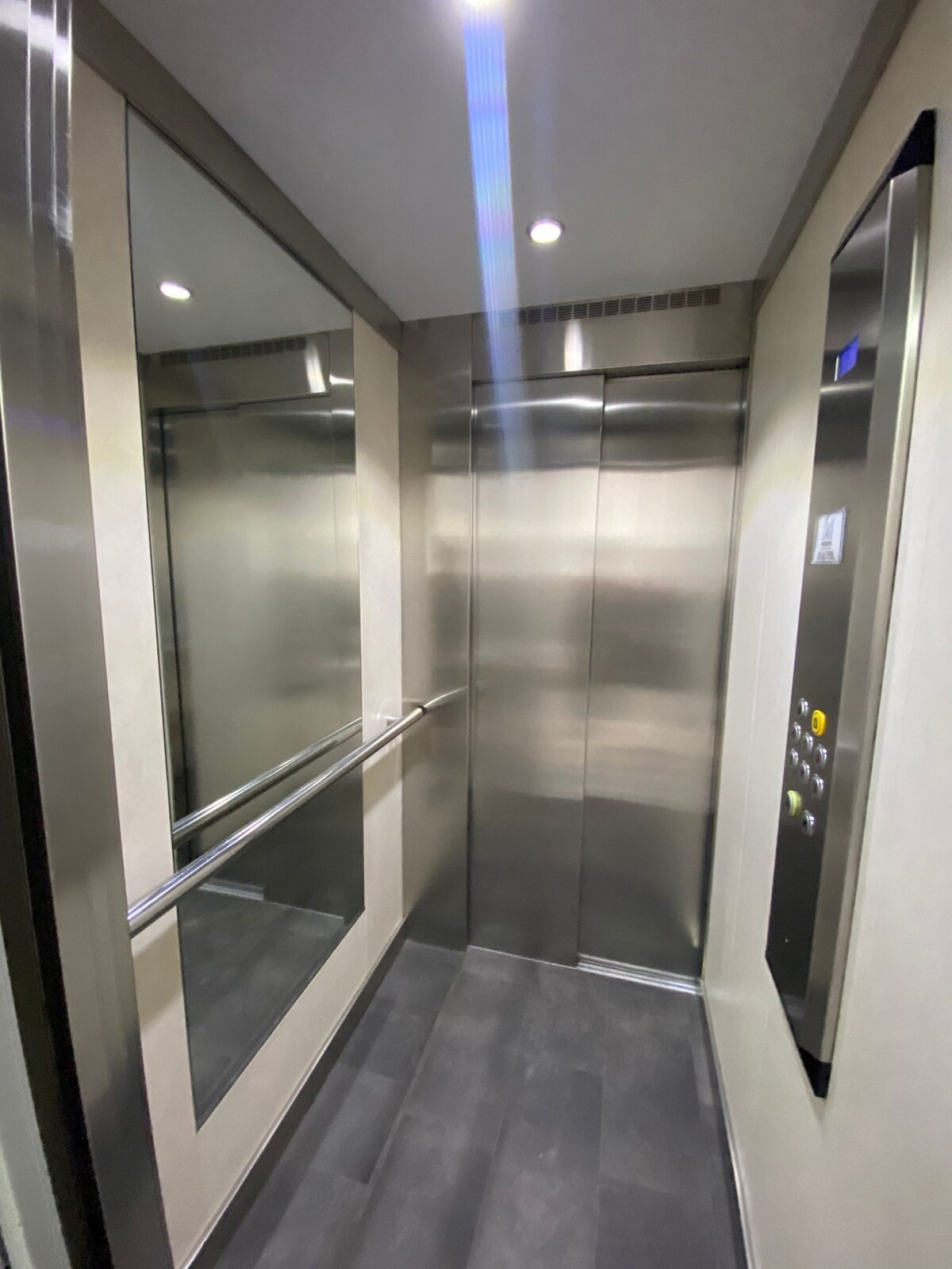 An elevator interior is displayed, featuring mirrored walls and a sleek stainless steel finish. The floor is covered with dark tiles, and a railing runs along one side. Soft overhead lighting creates a bright environment, with buttons for various floors visible near the entrance.