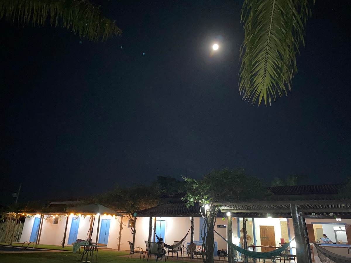 A serene night scene depicts a spacious house with illuminated windows. The moon casts a soft light over the exterior, highlighting surrounding greenery and gently swaying palm leaves. Hammocks are visible, inviting relaxation in the tranquil atmosphere.