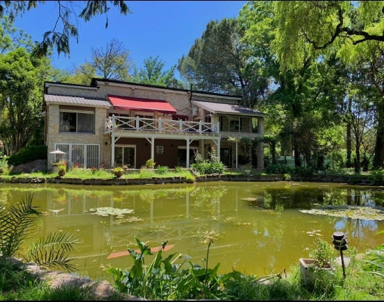A two-story house is positioned beside a serene, green-tinted lake, reflecting its surroundings. Large windows and a balcony overlook the water, while lush gardens enhance the natural setting. The peaceful ambiance is enriched by foliage and flowerbeds at the water's edge.