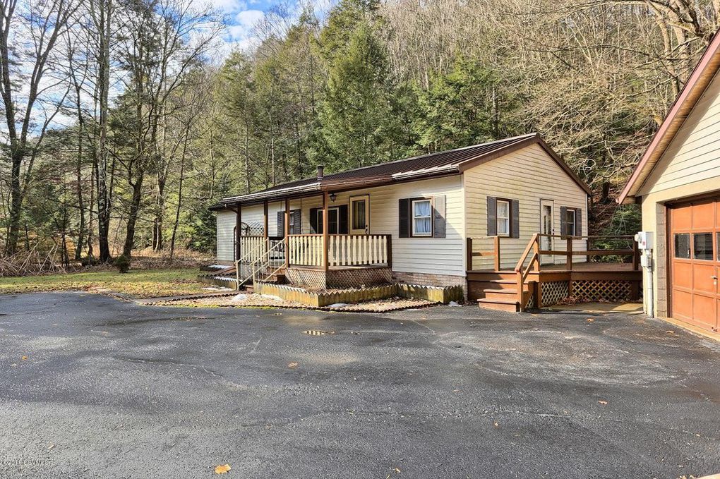 The exterior of the cottage is shown, featuring a wooden deck with steps leading to the entrance. Surrounding greenery includes tall trees and a well-maintained yard. A spacious driveway is visible, providing ample parking space.
