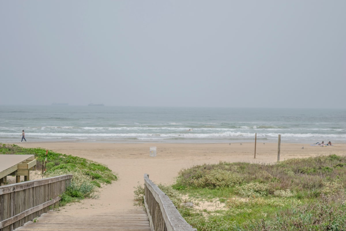 A sandy beach stretches into the distance, meeting the gentle waves of the ocean under a cloudy sky. A beachgoer is seen walking along the shoreline, while groups of people are scattered on the sand, engaged in various activities.