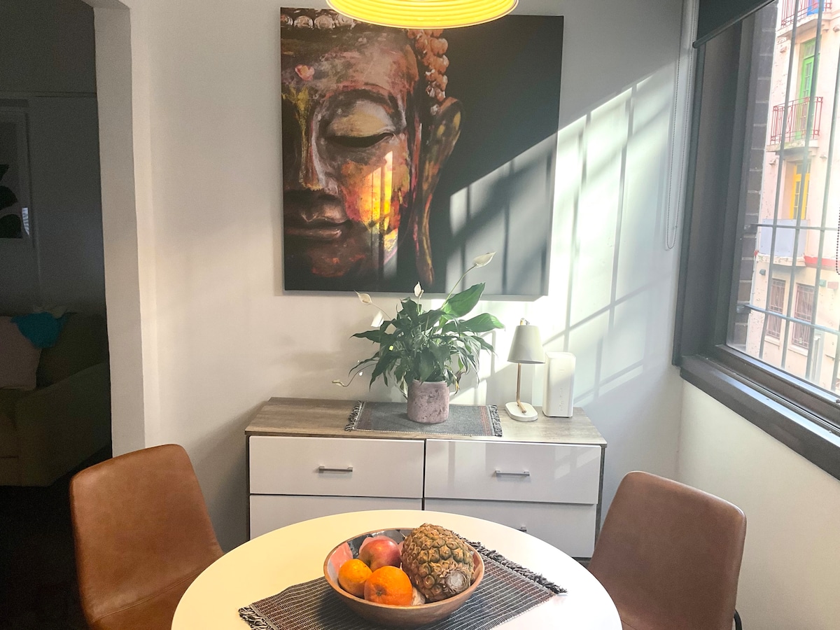 A dining area is presented with a circular table surrounded by two brown chairs. A decorative centerpiece of fruit and foliage is showcased, and a large artwork is displayed on the wall above the table. Natural light filters through a nearby window, enhancing the space.