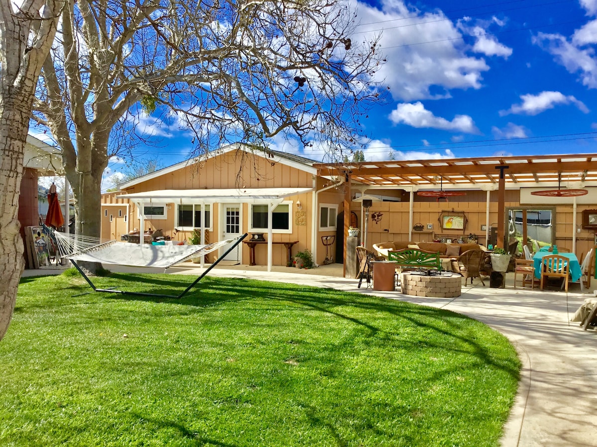 A spacious outdoor area features a well-maintained lawn and a hammock suspended between two trees. The studio is visible with a welcoming patio area furnished with seating and a dining table, set against a backdrop of a clear blue sky and fluffy white clouds.