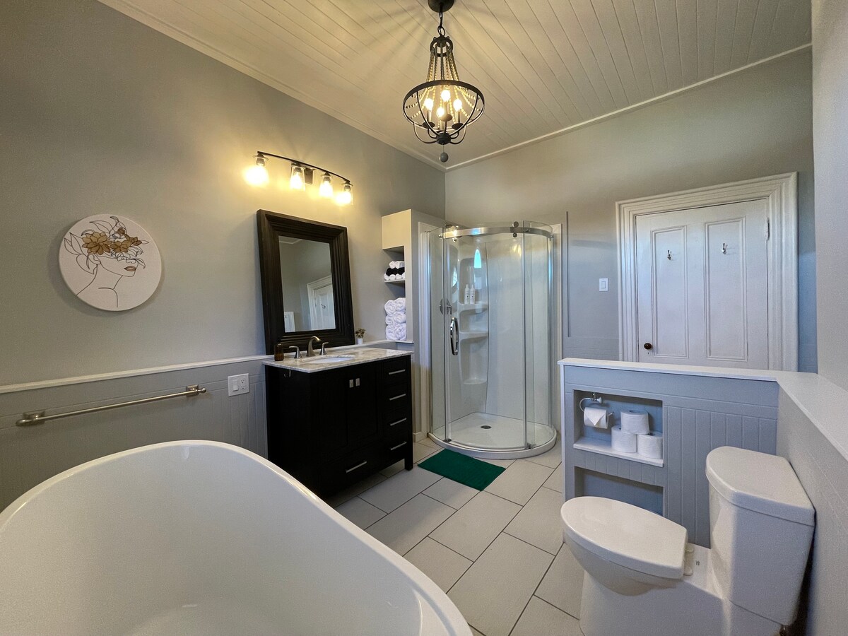 The spacious bathroom features a freestanding soaking tub and a modern shower enclosed in glass. A dark vanity with a sink is complemented by soft white towels stacked nearby. Neutral-colored walls and decorative lighting enhance the overall calming ambiance of the space.