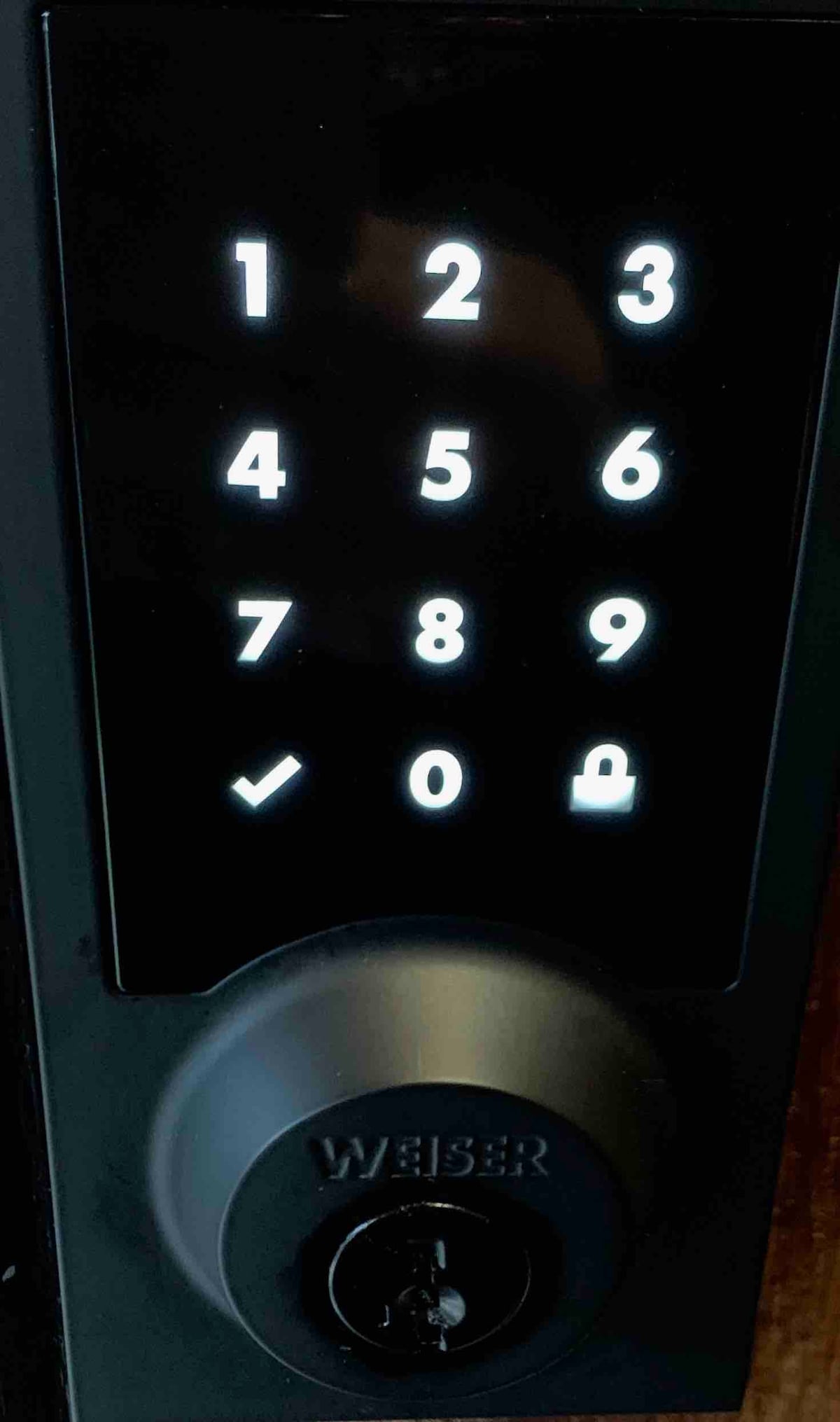 A close-up view of a digital keypad lock is displayed. The illuminated buttons feature numbers one through nine, with an additional zero and a checkmark symbol. A circular keyhole is also visible, presenting a modern security feature.