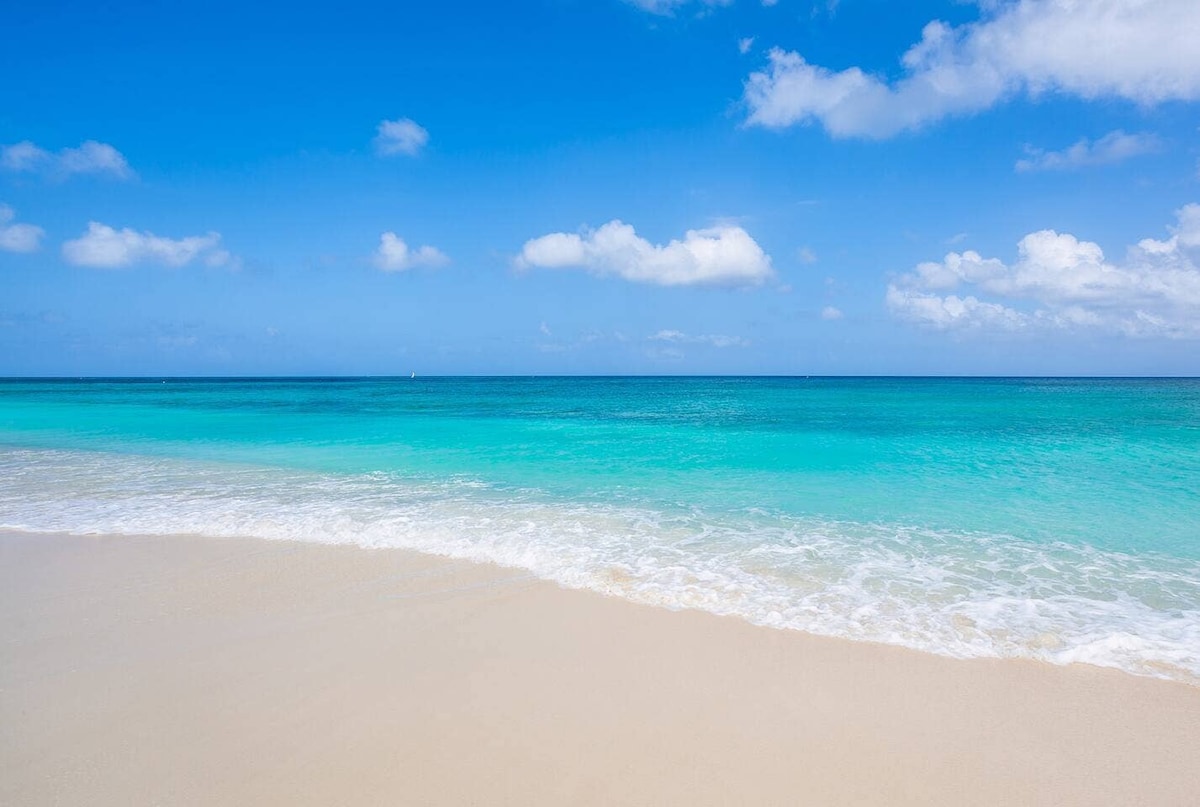 Gentle waves lap against softly curved sands, creating a serene shoreline. The vibrant blue water transitions to turquoise, meeting a sky dotted with light clouds, evoking a sense of tranquil outdoor space ideal for relaxation.