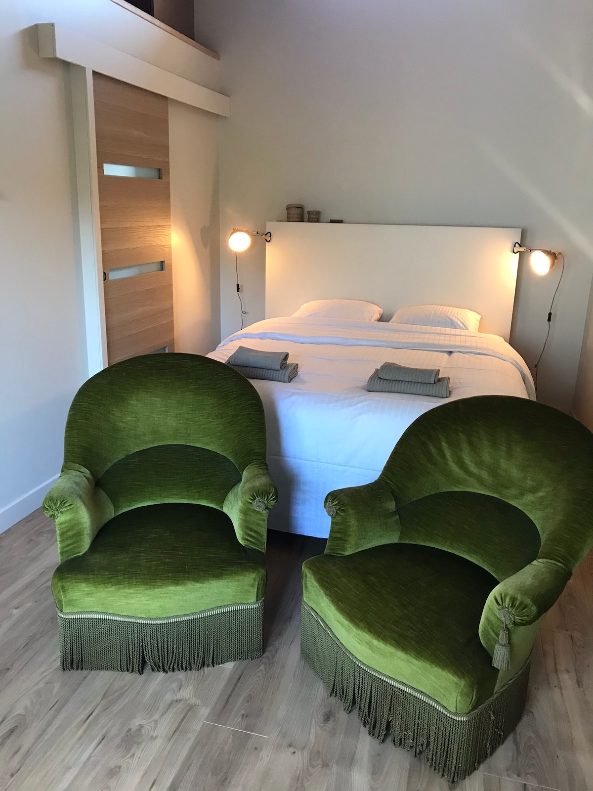 A sleeping area features a spacious white bed with neatly arranged linens. Two green upholstered armchairs with fringe detail are positioned at the foot of the bed. Soft lighting from wall sconces enhances the serene ambiance of the room.