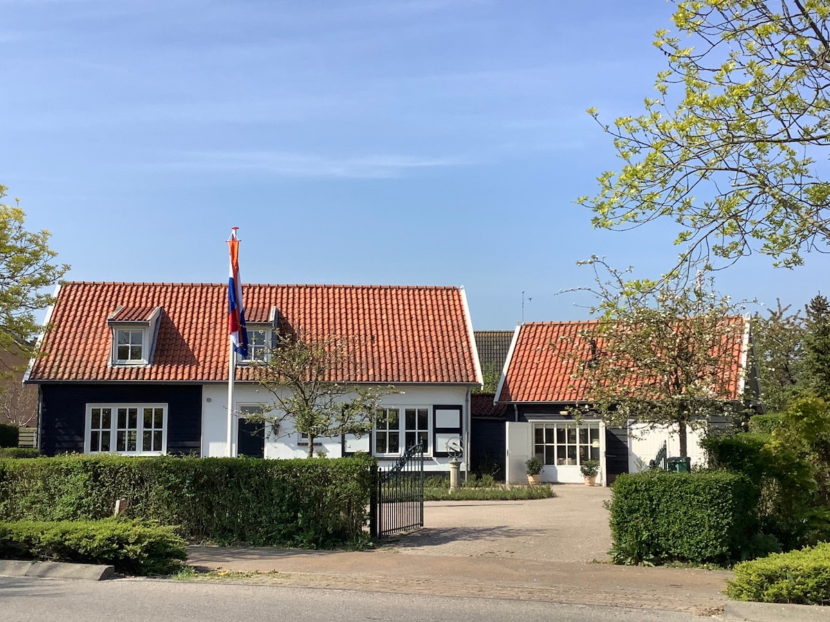 A charming guesthouse is depicted, featuring traditional Dutch architecture with a pitched red-tiled roof. The exterior is complemented by a neatly landscaped garden and hedges. A flag is displayed prominently, while the entrance is marked by a gated pathway leading to the front door.
