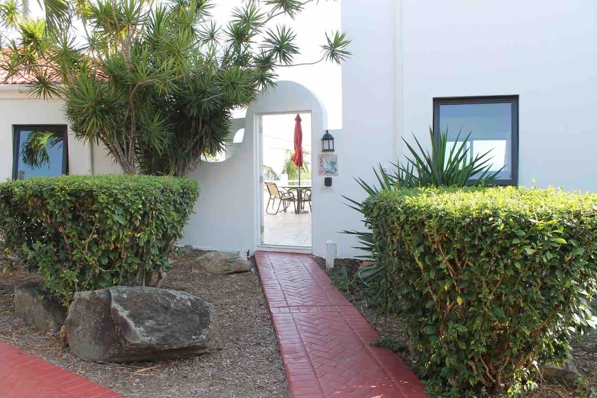 A landscaped pathway lined with lush greenery leads to an arched entrance. A red brick walkway is visible, complemented by carefully shaped shrubs and rocks. In the background, a patio area can be glimpsed through the doorway, where outdoor furniture and an umbrella are situated.