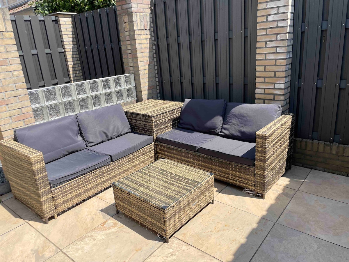 An outdoor seating area features a wicker sofa set with dark cushions. A low coffee table complements the seating, all situated on a tiled patio. The surrounding walls provide privacy, creating a serene environment for relaxation.
