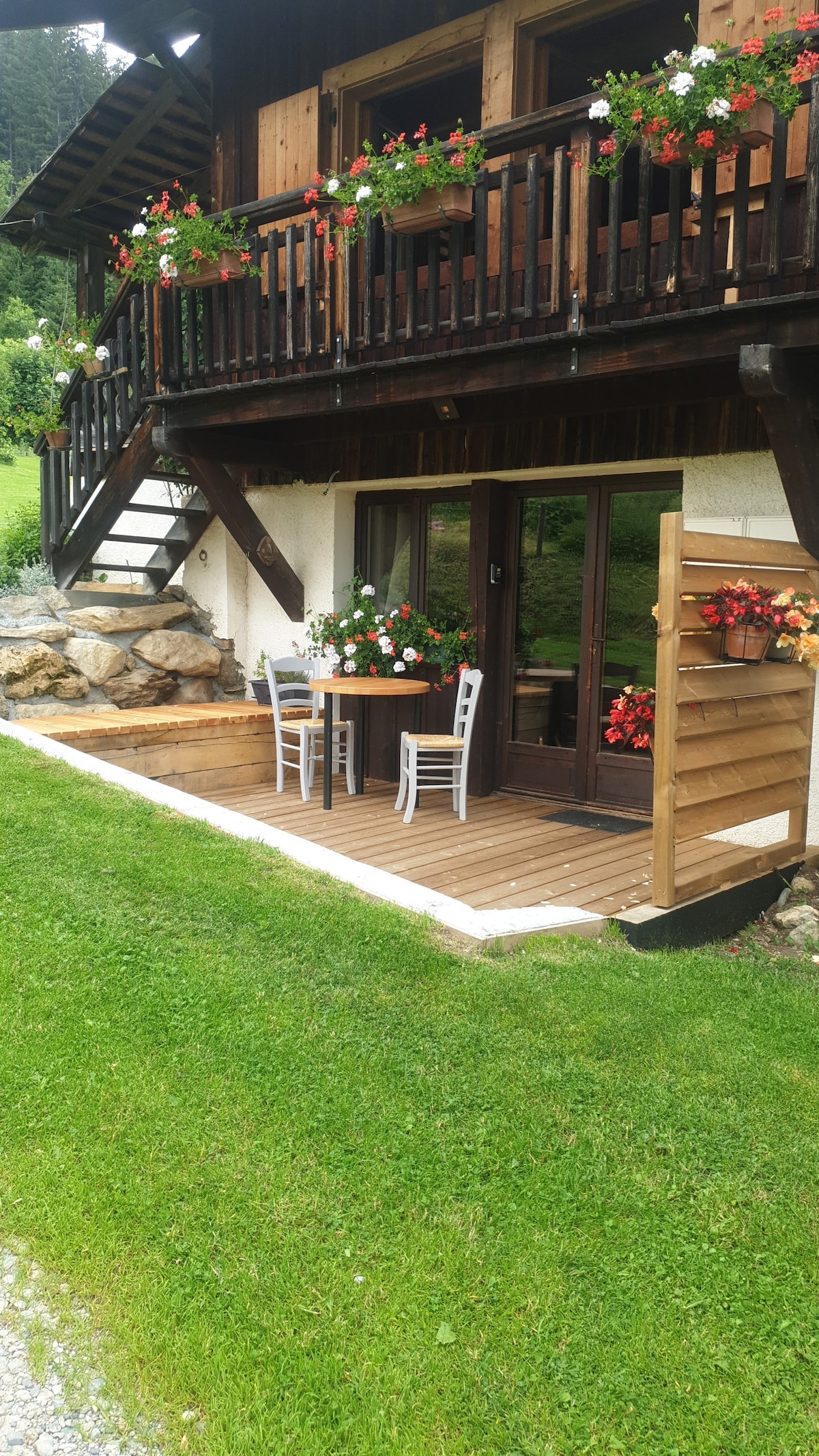 A private terrace is featured at the entrance, offering outdoor seating with a small table and two chairs. Flower boxes adorn the balcony above, providing a splash of color against the wooden exterior. Lush green grass surrounds the area, enhancing the peaceful ambiance.