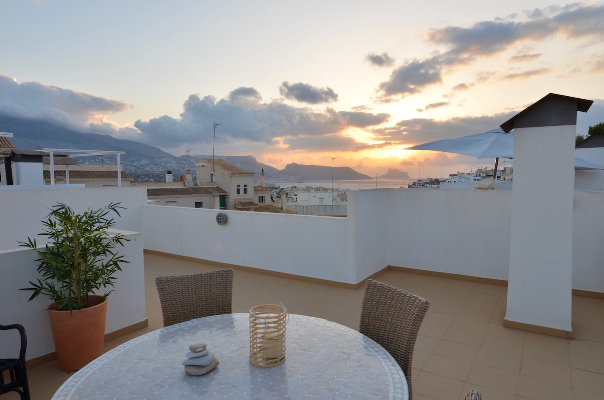 A rooftop terrace is presented with a round table surrounded by wicker chairs. A potted plant adds greenery, while a lantern sits on the table. The background showcases a vivid sunset over the distant mountains and sea.