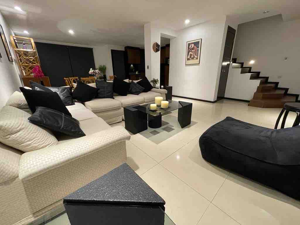 A spacious living area features a large 'L'-shaped sofa adorned with dark cushions. A central glass table is surrounded by smaller tables, while a puff adds additional seating. Stairs lead to the upper level, with a soft light illuminating the modern decor.