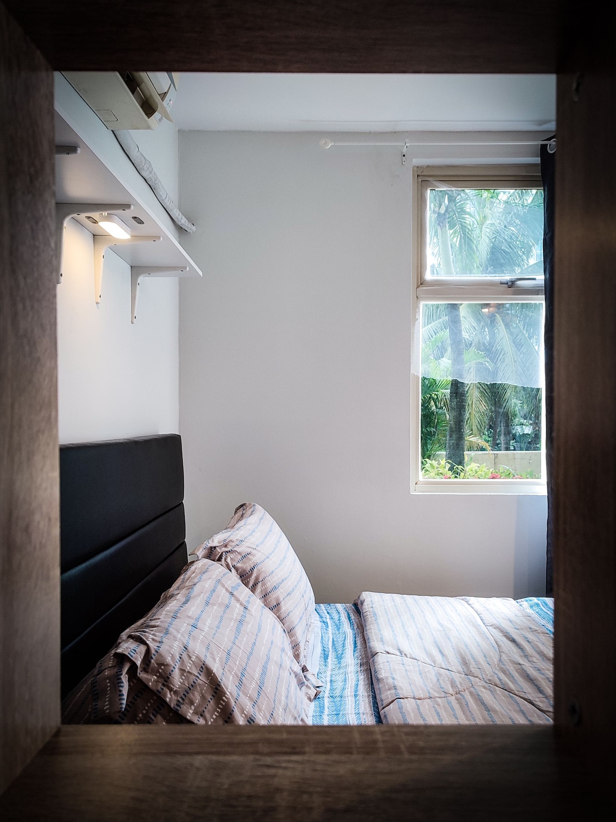 A cozy sleeping area is framed by a wooden border, featuring a spacious bed with lightly striped bedding. Natural light enters through a nearby window, revealing greenery outside and enhancing the serene environment.