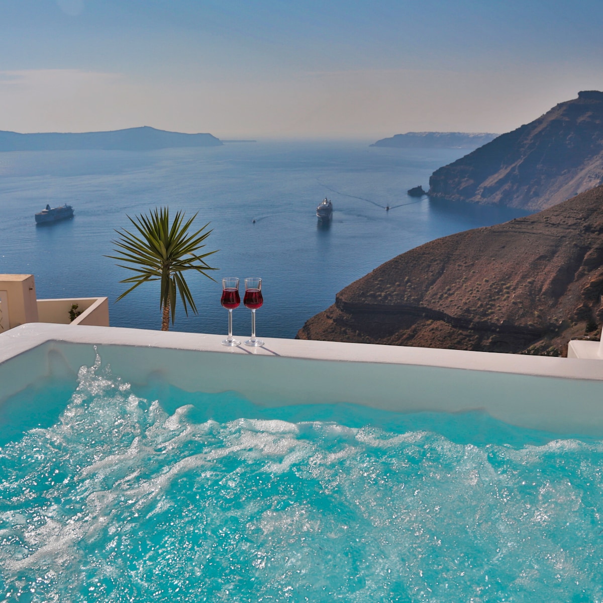 The image captures a heated Jacuzzi with clear blue water, positioned on a terrace overlooking the Aegean Sea. Two glasses of red wine sit at the edge, with a backdrop of ships navigating the calm waters and rugged cliffs rising from the shoreline.