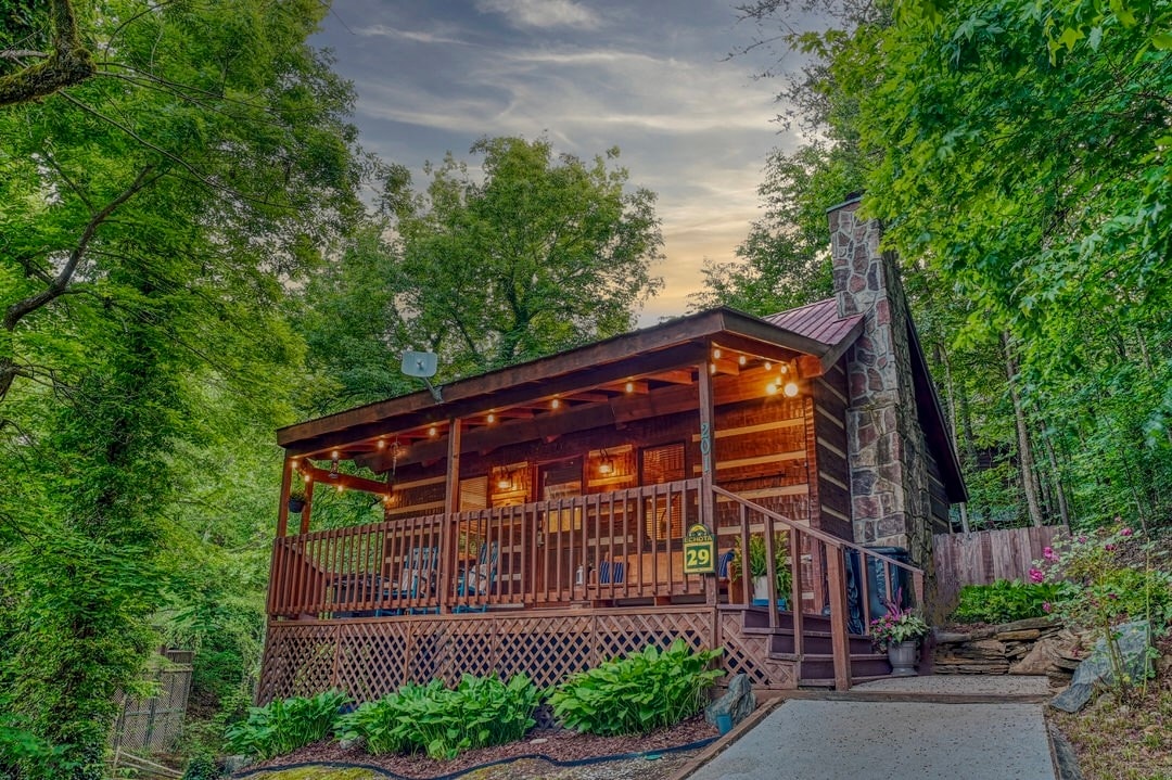 The cabin's front porch is surrounded by lush greenery, featuring comfortable seating arrangements. Stone details adorn the exterior, complemented by warm lighting, creating a welcoming presence nestled among the trees. A paved walkway leads to the entrance, highlighting the cabin's rustic charm.