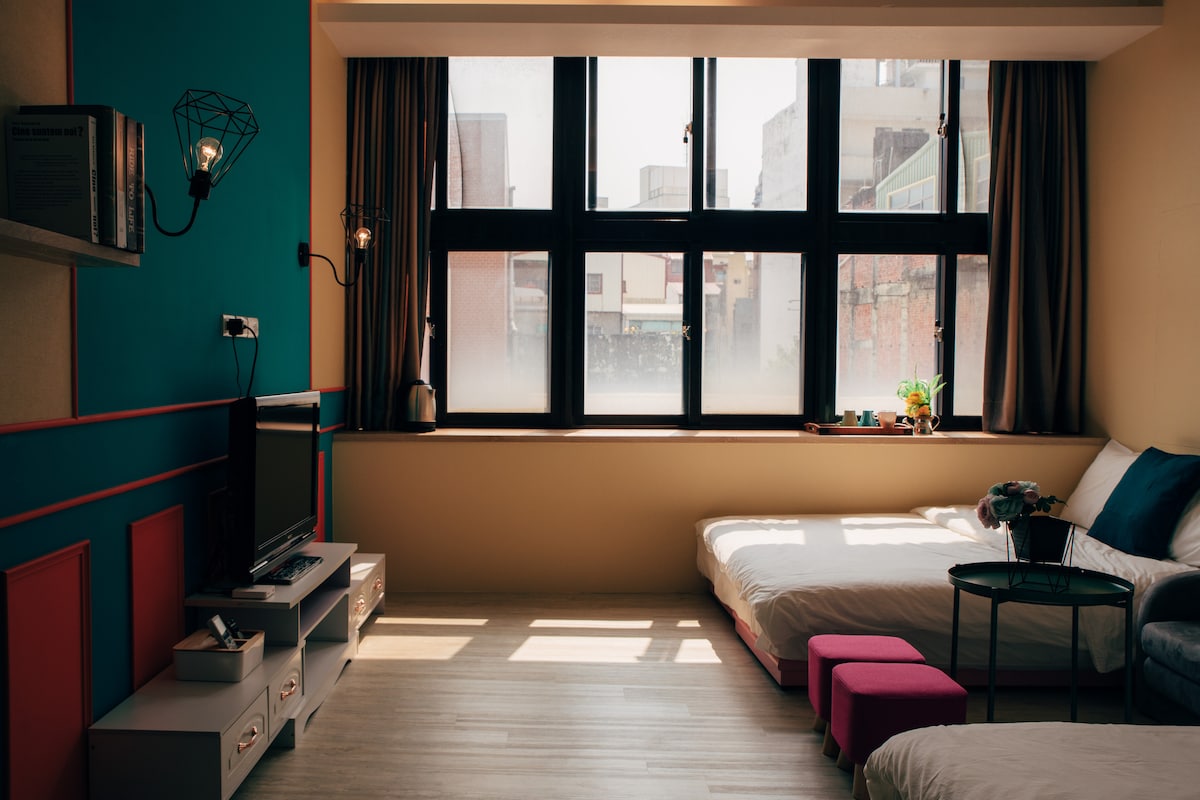 A bright room features a large window allowing natural light to fill the space, framed by light curtains. A spacious bed is complemented by a small side table and a cozy seating area. A television is placed on a low cabinet against the wall.