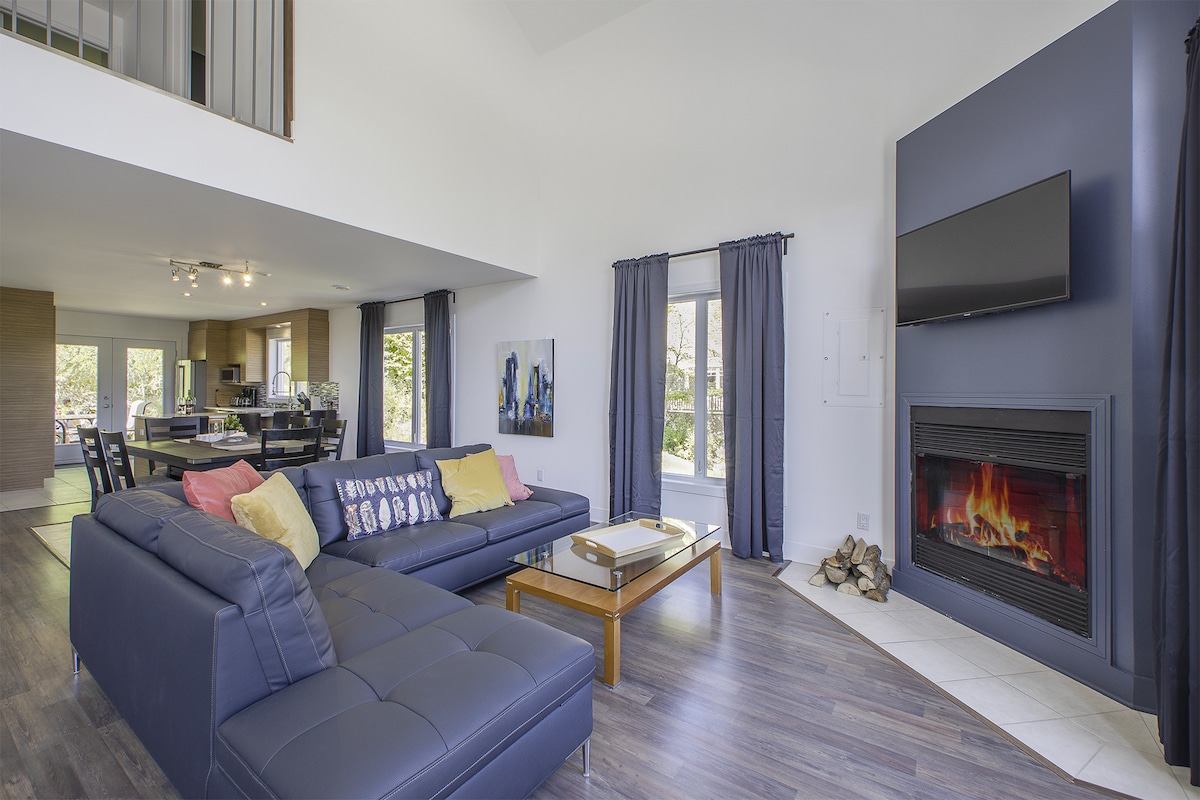 The spacious living area features a dark sectional sofa adorned with colorful throw pillows and a coffee table. A modern fireplace is situated beneath a flat-screen television. Large windows allow natural light to enhance the ambiance, while elegant curtains frame the view.