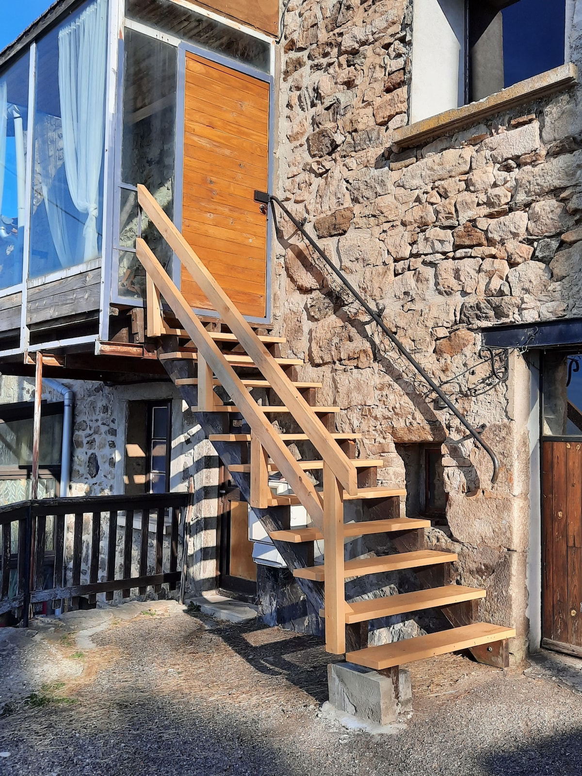 A set of wooden stairs leads to the entrance of the stone building, featuring a handrail for support. Natural light spills through a nearby window, and the entrance door is positioned at the top of the staircase, framed by the rustic stone facade.