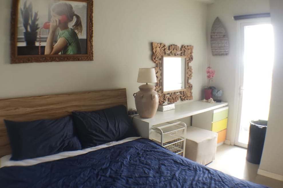 A cozy bedroom is presented with a wooden bed featuring navy bedding. A decorative mirror is mounted above a modern desk, accompanied by an ornate lamp and accent decor. Natural light enters through a nearby window, enhancing the serene atmosphere.