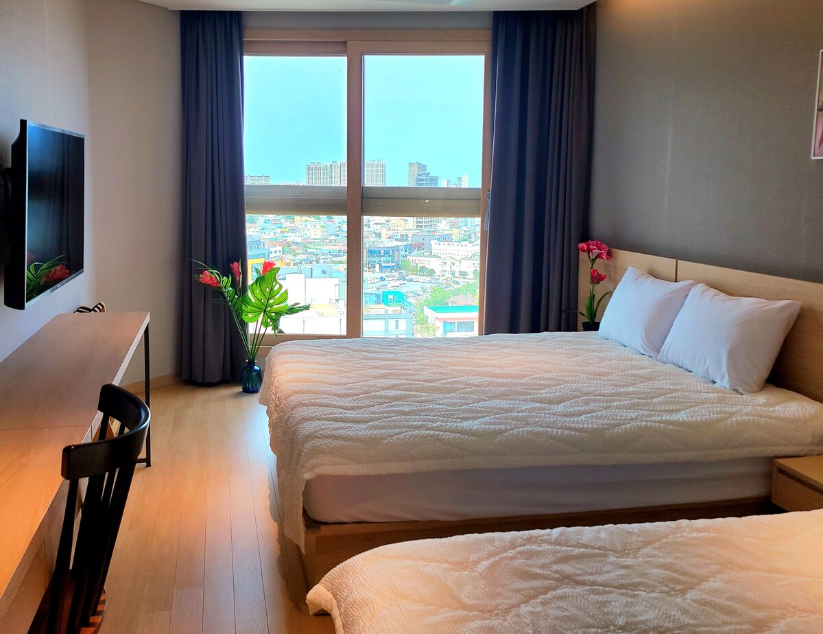 A bright sleeping area features two spacious beds with white bedding, complemented by a wooden desk and chair. Floor-to-ceiling windows provide scenic views of the surrounding area, allowing natural light to fill the room. Decorative plants add a touch of warmth.