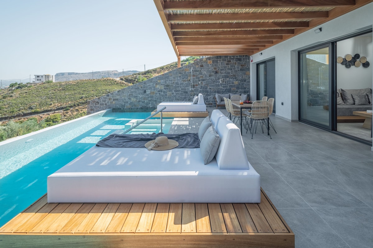The outdoor terrace features a heated infinity pool with stunning views. Two double daybeds are positioned on the wooden decking, complemented by a hanging chair. A dining area with a round table and chairs is also present, surrounded by natural stone walls and a pergola for shade.