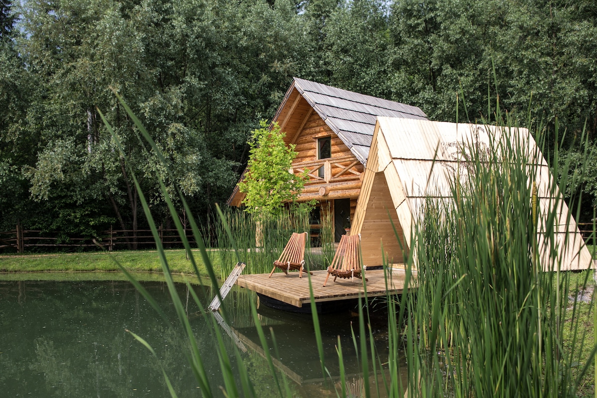 A wooden log house is situated beside a serene lake, surrounded by lush greenery. A deck extends into the water, featuring two wooden loungers. The structure's architectural style includes a gabled roof and large windows that integrate with the natural surroundings.