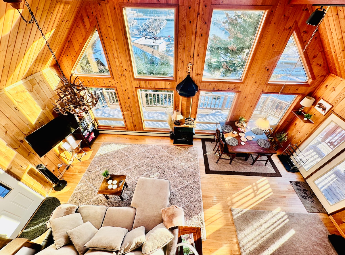 A spacious living area features large triangular windows, allowing natural light to fill the wooden interior. A comfortable seating arrangement is complemented by a coffee table, while a dining space with a view of the harbor is visible. Natural wood tones create a warm ambiance.