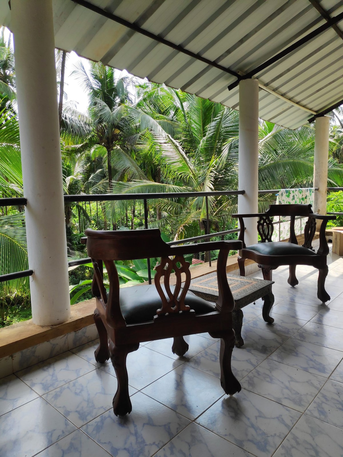 A balcony area is shown with two wooden chairs positioned beside a railing, overlooking lush greenery. The floor is tiled with a light marble pattern. The roof is supported by white pillars, creating an open space that invites natural light and views of the surrounding palm trees.